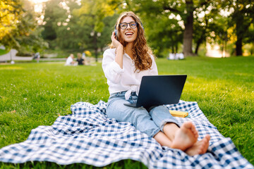 Portrait of a young woman sitting on a blanket with a laptop and wireless headphones on a lawn in a green park. Beautiful female freelancer enjoying music in headphones outdoors. Education concept.