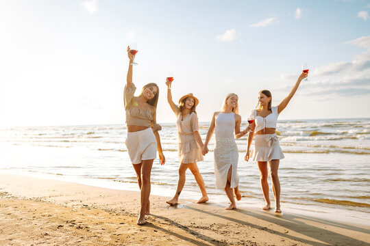 Four female friends having fun together on the seashore, drinking wine and relaxing on a mat. Beautiful women enjoying the sunset on the beach. Friendship and relaxation concept.