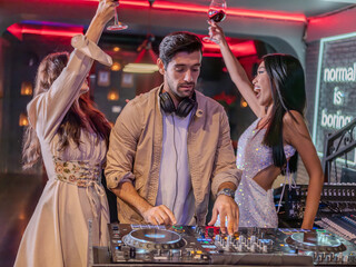 Group of stylish young adults enjoying a party in a nightclub with red neon lights. A DJ is mixing...