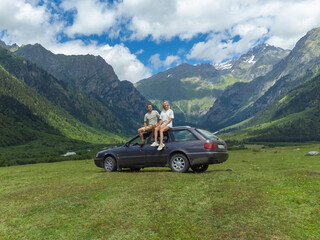 Adventurous couple sitting on the roof of their car, taking in the breathtaking panorama of a lush green valley nestled amidst towering mountains, enjoying a memorable road trip
