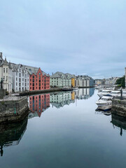 Colorful facades of art nouveau buildings with stunning reflections in the tranquil water of Brosundet canal and boats moored in a cloudy day in Alesund, Norway