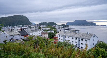 Scenic view of a residential neighborhood in Alesund, Norway, seen from Mount Aksla, with white houses, a coastal bridge, and mountainous islands in the background.