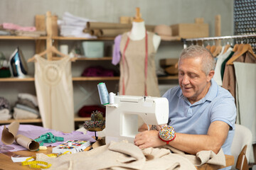 Mature man designer sews clothes using sewing machine. Worker creates wardrobe items, sews dresses...