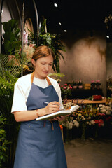 Middle-aged female florist in apron arranging calling on the smart phone. She is arranging...