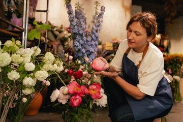 Middle-aged female florist in apron arranging fresh flowers for bouquet in the flower shop. Small...