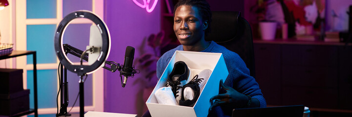 Young Black person holding sneaker box while sitting in front of microphone and ring light, showing...