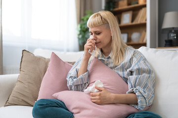 Ill upset young woman sitting on sofa covered with blanket freezing blowing running nose got fever...