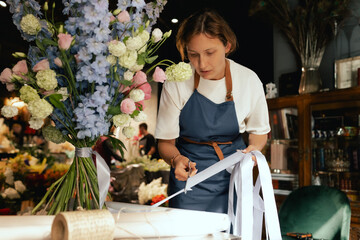 Middle-aged female florist in apron arranging fresh flowers for bouquet She ties a ribbon on the...