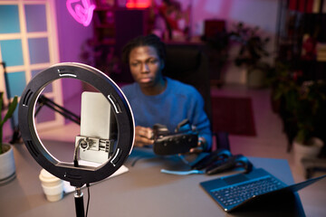 Individual sitting at desk with various technology devices creating online content, using smartphone, ring light, microphone in home studio environment