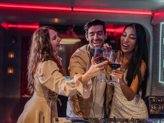 Group of stylish young adults enjoying a party in a nightclub with red neon lights. A DJ is mixing music while two women cheer with glasses of red wine, smiling and having fun.