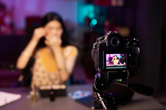 Woman filming makeup tutorial while being filmed by a professional camera in studio, with blurred purple neon background. Female influencer making content for vlog