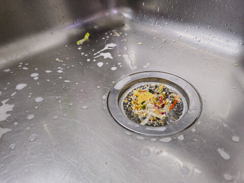 Close-up of a stainless steel kitchen sink with food scraps collected in a mesh drain strainer, surrounded by soap suds and water droplets.