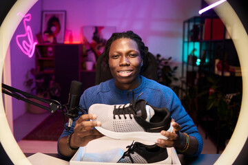Portrait of Black man showing running shoes in detailed studio setup with plants and neon lights, displaying content creating tools in professional environment