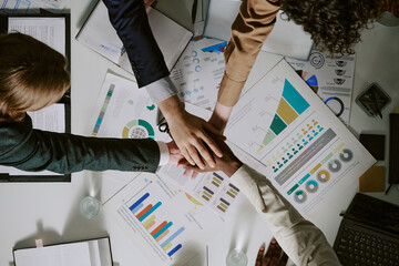 Multiracial business team joining hands over diverse charts and graphs, collaborating on business project in professional setting. Dynamic planning and teamwork by diverse colleagues