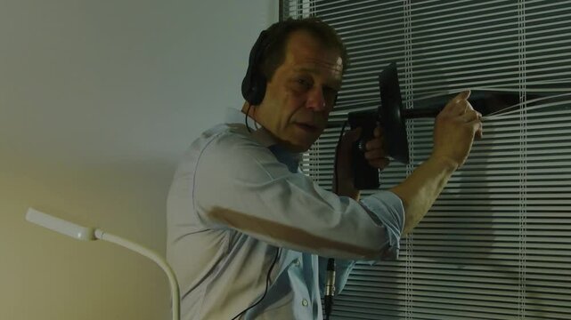 A man wearing headphones conducts covert surveillance through blinds using a directional microphone. The concept of espionage and covert investigation.