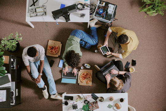 Group of diverse individuals sitting around table, working on various tech devices and eating pizza in modern office setting with greenery and tech equipment present