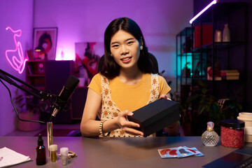 Woman smiling while holding a black beauty box during promotional livestream on social media. Neon lights and decor creating a cozy creative atmosphere