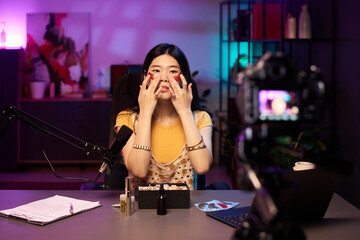 Portrait of an Asian woman applying eye patches in studio during vlogging, using modern gadgets surrounded by various cosmetics and lighting equipment