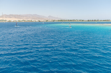 Expansive Red Sea view under bright daylight, water sparkling vividly. Mountains offer rugged backdrop beyond shoreline dotted with greenery, creating serene coastal scene