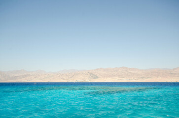 Crystal-clear turquoise waters stretch across Red Sea with distant arid mountains under cloudless sky. Tranquil seascape offers vibrant contrast of sea and land