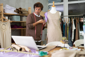 Fototapeta premium Man seamstress communicates with customer via video call work with mannequin. Contractor clarifies details with customer, discusses work issues using laptop device and remote technology.