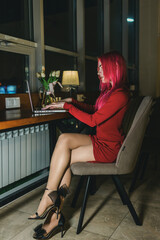 A girl with red hair and a red dress is sitting at a laptop in a cafe
