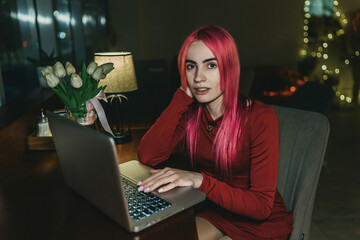 A girl with red hair and a red dress is sitting at a laptop in a cafe