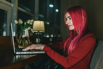 A girl with red hair and a red dress is sitting at a laptop in a cafe