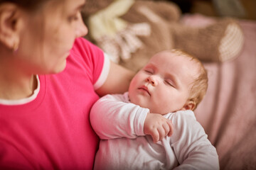Young woman with braided hair gently cradles sleeping baby girl in pink and beige room. Warm lighting and soft textures create calm atmosphere