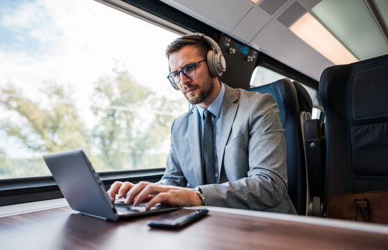 Young businessman wearing headphones and suit is making a video conference on his laptop while enjoying a cup of coffee during his train journey