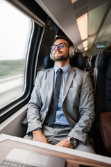 Young businessman wearing suit and tie is sitting on a train next to the window, listening to music with headphones and enjoying his comfortable trip