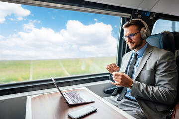 Young businessman wearing headphones and suit is making a video conference on his laptop while enjoying a cup of coffee during his train journey