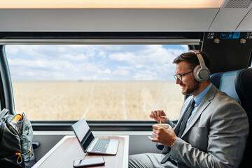Young businessman wearing headphones and suit is making a video conference on his laptop while enjoying a cup of coffee during his train journey