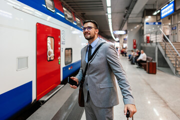 Elegant businessman waiting for a train at a bustling railway station, holding a mobile phone while pulling a trolley suitcase and gazing thoughtfully into the distance