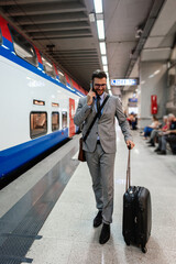 Young businessman dressed in a stylish suit, carrying a suitcase, walking through a bustling train station while smiling and talking with someone on the phone.