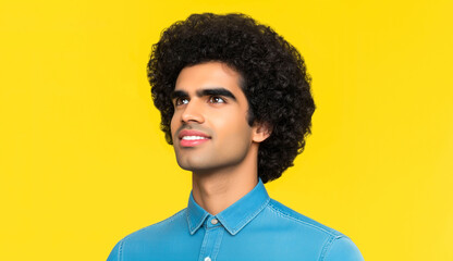 Fototapeta premium Portrait of a happy young Indian man with curly hair wearing a denim shirt, laughing and looking away over a yellow background. Concept with copy space.