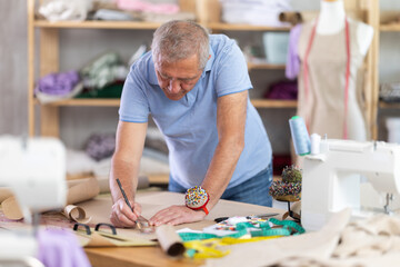 Fototapeta premium Tailor stands near the table and draws patterns of clothes with a pencil on paper. Work in the sewing workshop
