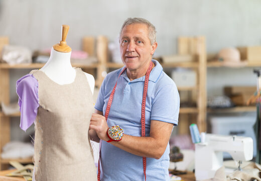 Elderly male tailor adjusting clothes on mannequin in sewing workshop