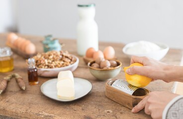 Hands gently grate bright lemon zest over a wooden box grater, surrounded by eggs, nuts, and milk, showcasing a warm, inviting kitchen atmosphere perfect for baking