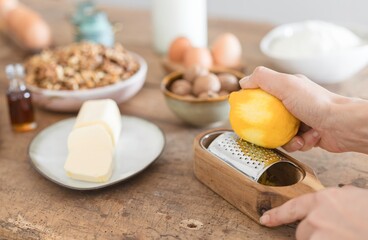 A hand expertly grates a bright yellow lemon over a wooden zester, infusing zest into a bowl filled with baking ingredients. The warm kitchen setting enhances the culinary experience