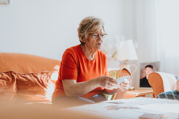 An elderly woman wearing an orange shirt spends a peaceful moment playing cards at the table in a warmly lit living room.