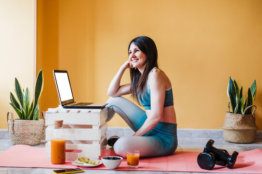 Thoughtful woman looking away while sitting with laptop on exercise mat at home