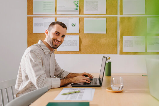 Businessman working on a laptop in an office with a corkboard and documents