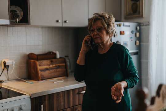 An elderly woman in glasses holds a phone to her ear while standing in a kitchen, appearing in conversation with a concerned expression.