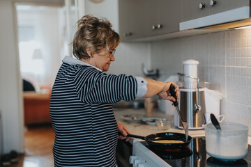 An elderly woman preparing food in her kitchen. She uses a spatula and non-stick frying pan to make...