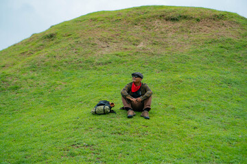 An adventurer is resting by sitting on a stretch of green grass. The Asian man is enjoying the tranquility of nature by resting on a green hill that turns out to be a buried Buddhist temple.