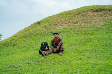 An adventurer is resting by sitting on a stretch of green grass. The Asian man is enjoying the tranquility of nature by resting on a green hill that turns out to be a buried Buddhist temple.