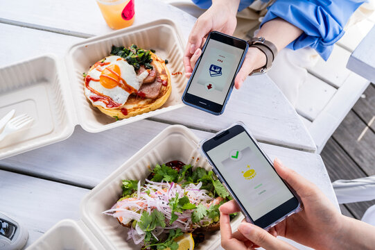 Two women complete a mobile payment transaction at an outdoor seaside café, each holding a smartphone displaying fintech apps. - Powered by Adobe