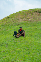 An adventurer is resting by sitting on a stretch of green grass. The Asian man is enjoying the tranquility of nature by resting on a green hill that turns out to be a buried Buddhist temple.