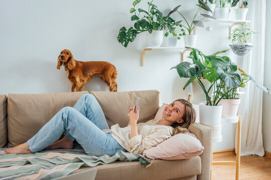 Cheerful woman holding smart phone while lying on sofa by dog in living room